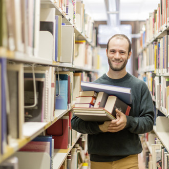 Ein Student steht in der Bibliothek im Gang und hat Bücher auf dem Arm.