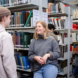 Zwei sich unterhaltende Studierende und ein lesender Studierender in der Bibliothek.