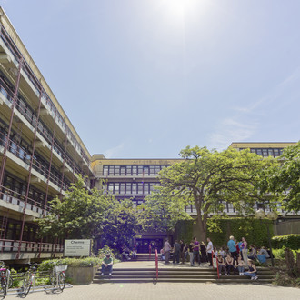 Chemiegebäude: Ein großes, breites, graues Gebäude mit Fensterfronten und Balkonen vor den Fenstern, die mit roten Metallstangen abgegrenzt werden. In der Mitte eine Treppe, auf der Menschen sitzen und links vor der Treppe Fahrräder in Fahrradständern.