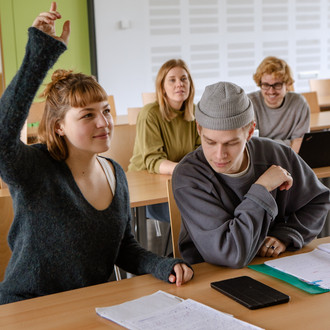 Studierende im Seminarraum sitzen an den Tischen. Eine Person meldet sich. 