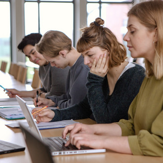 Studierende im Seminarraum sitzen in einer Tischreihe vor ihren Laptops. 