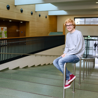 Student sitzt auf einem Geländer an der Treppe und lächelt in die Kamera. 