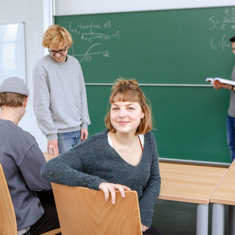 Zwei Studierende sitzen im Vordergrund im Seminarraum. Ein Studierender steht mit einem Dozierenden im Hintergrund an der Tafel.