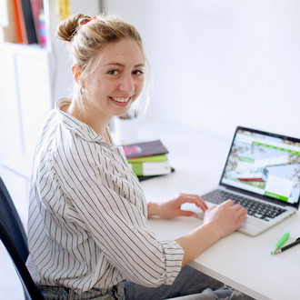 Am Schreibtisch sitzende Studentin tippt auf dem Laptop, daneben liegen Bücher und Stifte.