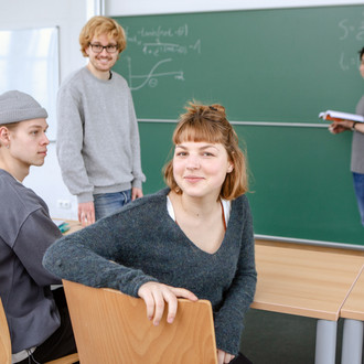 Zwei Studierende im Seminarraum im Vordergrund sitzen. Ein Studierender steht mit dem Dozierenden an der Tafel.