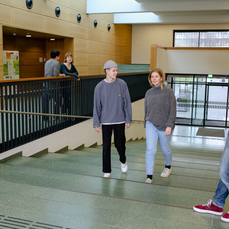 Studierende im Seminargebäude 1, im Vordergrund steht ein Student am Geländer und schaut auf zwei andere Studierende, die die Treppe hochgehen.