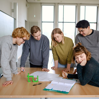 Fünf Studierende im Seminarraum stehen um einen Tisch herum und sehen auf ein Dokument