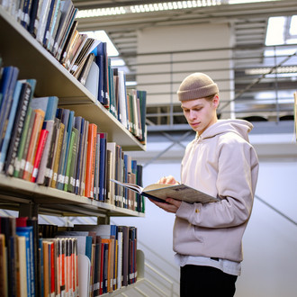 Studierender steht vor Bücherregal und liest in einem Buch.