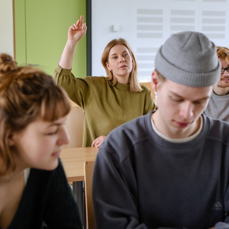 Studierende im Seminarraum sitzen an den Tischen. Eine Person meldet sich. 