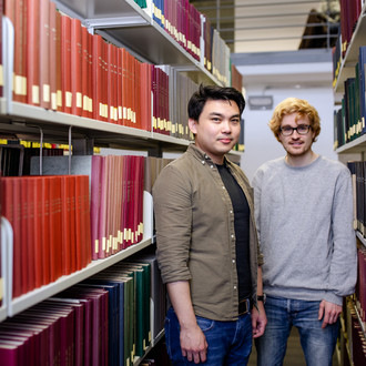 Zwei Studierende stehen zwischen zwei Bücherregalen in der Bibliothek.