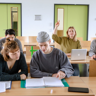 Studierende im Seminarraum sitzen an den Tischen. Eine Person meldet sich. 