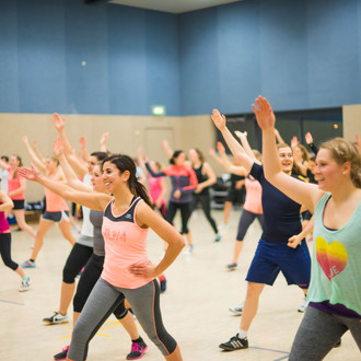 Teilnehmerinnen und Teilnehmer eines Fitness-Kurses in einer großen Sporthalle