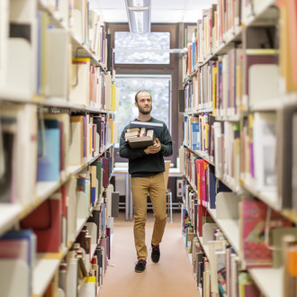 Studierender läuft an den Bücherregalen in der Bibliothek entlang.