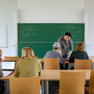 Vier Studierende im Seminarraum sitzen und arbeiten zusammen mit dem Dozierenden im intergrund.