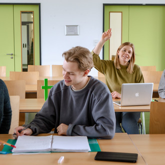 Studierende im Seminarraum sitzen an den Tischen. Eine Person meldet sich. 