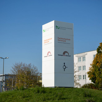 Hinweisschild auf einem Kreisverkehr, im Hintergrund ein Gebäude und Bäume.