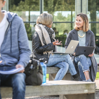 Zwei Studentinnen unterhalten sich draußen. Sie haben Laptops auf dem Schoß.