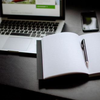 Eingeschalteter Laptop, Smartphone und Notizbuch mit Stift auf einem Tisch liegend.