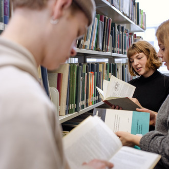 Drei Studierende stehen zwischen zwei Bücherregalen und lesen.