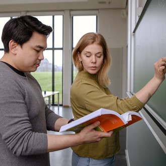 Zwei Studierende stehen an der Tafel, sie schreibt aus dem Buch ab, welches er hält