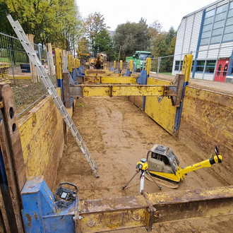 An einer Straße auf dem Campus ist ein langer rechteckiger Aushub, in dem Baustellengeräte und gelbe Metallstützen zu sehen sind. Abgesichert wird die Baustelle auf beiden Straßenseiten durch einen Metallzaun.