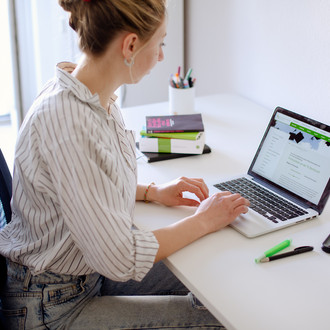 Am Schreibtisch sitzende Studentin tippt auf dem Laptop, daneben liegen Bücher und Stifte.