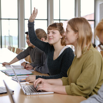 Studierende im Seminarraum sitzen in einer Tischreihe vor ihren Laptops. Eine Studierende meldet sich.
