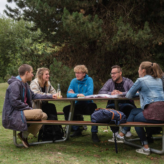 Sechs Studierende sitzen an einer Sitzgruppe auf der Wiese mit Bäumen im Hintergrund. 