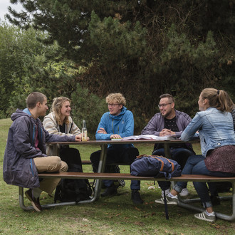 Sechs Studierende sitzen an einer Sitzgruppe auf der Wiese mit Bäumen im Hintergrund. 