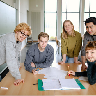 Fünf Studierende im Seminarraum stehen um einen Tisch herum und arbeiten gemeinsam