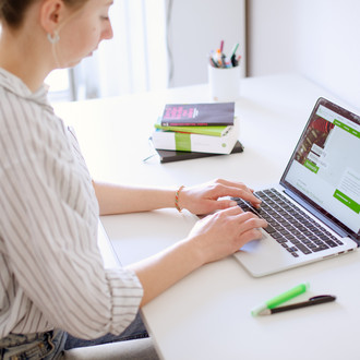 Am Schreibtisch sitzende Studentin tippt auf dem Laptop, daneben liegen Bücher und Stifte.