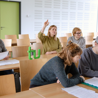 Studierende im Seminarraum sitzen an den Tischen. Eine Person meldet sich. 