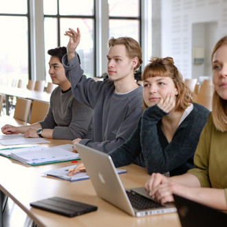 Studierende im Seminarraum sitzen in einer Tischreihe vor ihren Laptops. 