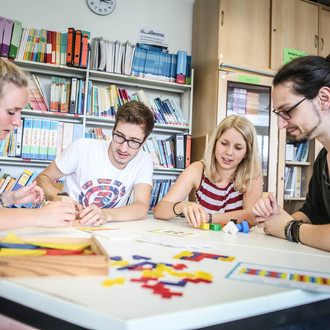 Vier Personen sitzen an einem Tisch und beschäftigen sich mit Bausteinen, die auf dem Tisch liegen. Im Hintergund steht ein Regal voller Bücher und Ordner und ein Schrank.