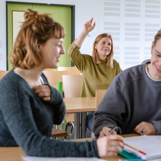 Studierende im Seminarraum sitzen an den Tischen. Eine Person meldet sich. 