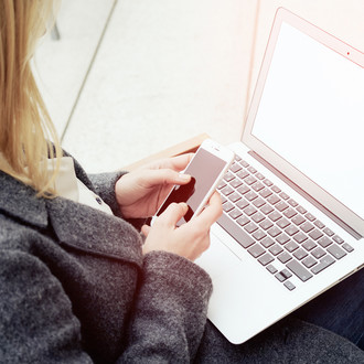 Frau mit Laptop auf dem Schoß, hält ein Smartphone in den Händen.