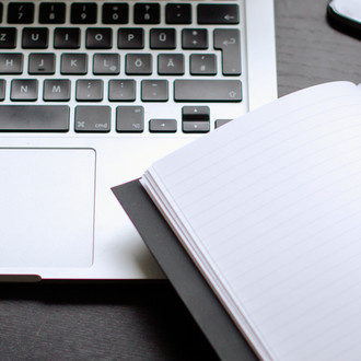 Laptop und Notizbuch mit Stift auf einem Tisch liegend.