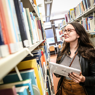 Studierende steht mit einem Tablet in der linken Hand an den Bücherregalen in der Bibliothek und sucht ein Buch.