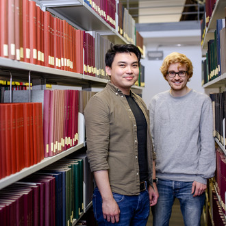 Zwei Studierende stehen zwischen zwei Bücherregalen in der Bibliothek. 