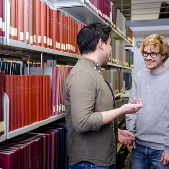 Zwei männliche Studierende unterhalten sich zwischen Regalreihen in der Bibliothek.