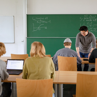 Vier Studierende sitzen im Seminarraum zur Tafel gewandt. Der Dozierende erklärt etwas.