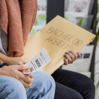 Zwei Personen sitzen nebeneinander. Man sieht einen Ausschnitt von Knie bis Brust. Die hintere Person hat einen großen Umschlag mit der Aufschrift „Bachelor-Arbeit“ in der Hand, die vordere Person hat einen kleinen weißen Zettel mit einer Wartenummer, einem Code und dem TU-Logo in der Hand.