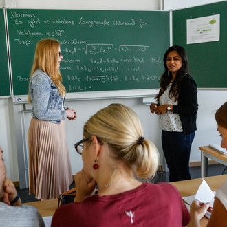 Im Hintergund stehen zwei Personen vor einer Tafel, auf die mit Kreide Formeln geschrieben sind. Im Vordegrund sieht man drei Personen von hinten, die an einem Tisch sitzen.