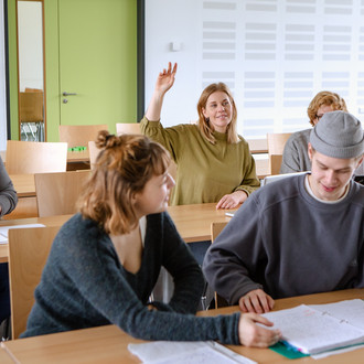 Studierende im Seminarraum sitzen an den Tischen. Eine Person meldet sich. 