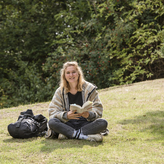 Eine Studentin sitzt im Schneidersitz auf einer Wiese mit einem Buch in der Hand.