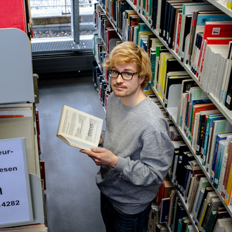 Studierender steht lesend vor einem Bücherregal in der Bibliothek.