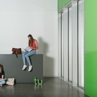 Eine Studentin sitzt auf dem Boden und liest, eine andere auf einer Theke und liest. Rechts im Bild ein großes Fenster.