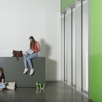 Eine Studentin sitzt auf dem Boden und liest, eine andere auf einer Theke und liest. Rechts im Bild ein großes Fenster.