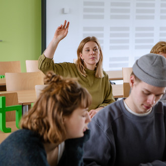 Studierende im Seminarraum sitzen an den Tischen. Eine Person meldet sich. 