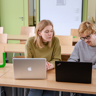 Zwei Studierende im Seminarraum sitzen vor ihren Laptops. Daneben das TU Logo.